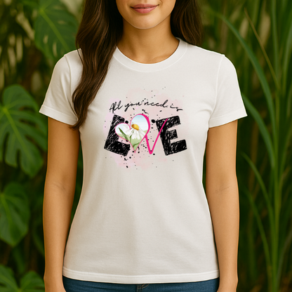 Woman wearing a white “All You Need Is Love” graphic T-shirt with daisy and heart design, standing among green plants; cropped from chin down, showcasing untucked boyfriend-fit shirt and top of blue jeans.