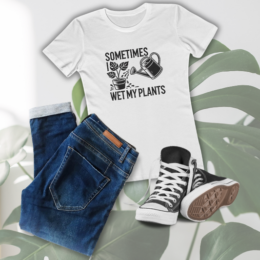 Flat lay of a white “Sometimes I Wet My Plants” graphic T-shirt in a women’s boyfriend fit, styled with cuffed blue jeans and black high-top sneakers, laid on a tropical monstera leaf background.