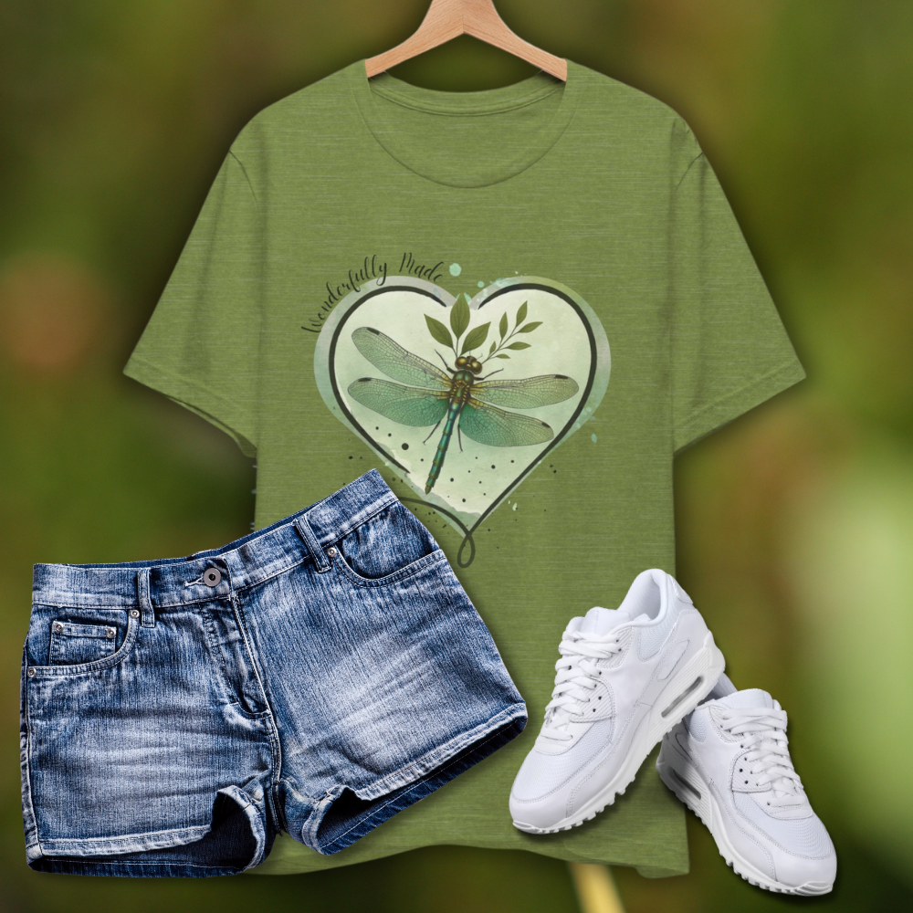 Olive green “Wonderfully Made” dragonfly T-shirt displayed with denim shorts and white sneakers on a soft green nature-inspired background; features a heart-shaped watercolor design with a botanical dragonfly and Christian message.
